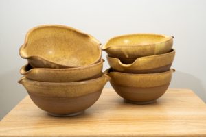 the photograph shows two stacks of three cassoulet bowls resting on a small light colored wooden table. the stack on the left side of the photograph is slightly closer to the camera then the stack on the right. both stacks contain 3 cassoulet bowls glazed in yellow salt. each bowl has the glaze cover the inside completely and the outside upper lip. the outside bottom of each bowl is unglazed and shows off the natural color of the clay (reddish brown). the background is a plain white wall. the photograph is lit with white light.