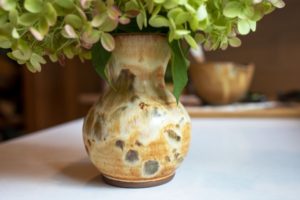 a horizontally framed photograph shows a close up of a clay coyote small vase glazed in feather. the vase has a bouquet of flowers in it, the flowers are mostly cut off out of frame, but the bit visible is bright green with hints of pink. the bouquet of flowers is larger then the vase. the vase is sitting on a white kitchen counter top. the photograph was taken at counter top height, giving the vase a "head on" view. in the background, to the right and further down the kitchen counter top is a clay coyote whiskey bowl glazed in joes blue. it is out of focus.