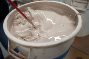 a horizontally framed photograph shows a close up of the top of a clay coyote glazing bucket. the surface of the glaze is turbulent, as a red mixing attachment is currently mixing the glaze. the photograph is close, close enough that not all of the top of the bucket is visible. the upper middle of the buckets rim is cut out of frame, with the rest of the rim being visible. the bucket is white.