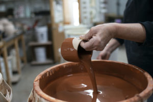 a horizontally framed photograph shows the process of glazing a clay coyote yunomi wine cup. the potter is standing on the right side of the photograph, with the glaze bucket taking up the lower right portion of the photograph. the potter is using one hand to hold the yunomi at an angle, pouring out the excess glaze from the inside. the outside of the yunomi shows it has just been dipped into the brown colored glaze. the area directly around the potters fingers remain unglazed. the potter is wearing a black long sleeve t-shirt with the sleeves pulled up to mid forearm. the background is out of focus but shows the workspace.