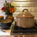 a horizontally framed picture. a clay coyote dutch oven is sitting on the iron grate of a stove top gas burner range. The burner is lit and there is a small blue flame visible underneath the dutch oven. The dutch oven is in coyote grey with darker speckles through out the glaze. The lid is on, and the handles are pointing to the left and right sides of the frame. To the left of the dutch ovens are two stacks of clay coyote chili bowls. the nearest stack of bowls is 3 high with dark glazes visible, and the top bowl is in mint chip glaze. The second stack of bowls, slightly behind the first and to the left is 5 high with 2 joes blue, 1 mint chip and a yellow salt glaze on the top. the bottom bowl is hard to tell glaze. Behind the stack of first bowls is a clay coyote large vase in joes blue, filled with a fall bouquet with orange, yellow and red flowers