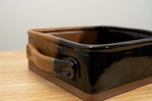 the photograph shows a clay coyote baking dish glazed in mocha swirl resting on a small light colored wooden table. the brown part of the mocha swirl pattern is in the back left corner of the dish. the background is a white wall. the photograph is lit with white light.