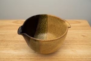 the photograph shows a high angle view of a clay coyote mixing bowl glazed in tiger. the mixing bowl is resting on a small light colored wooden table. the background is a plain white wall.