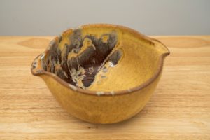 the photograph shows a high angle view of a clay coyote mixing bowl glazed in tequila sunrise. the angle allows the viewer to see the tequila sunrise pattern of the glaze inside the mixing bowl. the mixing bowl is resting on a small light colored wooden table. the background is a plain white wall.