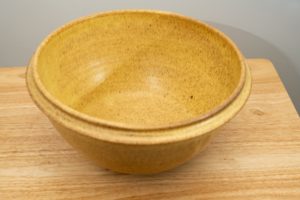 the photograph shows a high angle view of a clay coyote deep salad bowl glazed in yellow salt. the angle is high enough to allow the viewer to see the glaze details in the bowl. the deep salad bowl is resting on a small light colored wooden table. the background is a white wall.