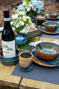 the photograph shows a close up of a tablescape made with clay coyote pottery. everything is on a light colored wooden table. starting from the left of the photograph working right there are: a clay coyote wine coaster, a yunomi, (both glazed in joes blue) a small vase glazed in zappa, a baking dish glazed in joes blue, a sandwich plate glazed in joes blue, with a joes blue soup and chili bowl resting on top of it and finally in the upper right corner, there is another sandwich plate and soup/chili bowl combo both done in joes blue. there are small branches of ferns decoratively laid on the table. each place setting has a blue placemat underneath the sandwich plate. the small zappa vase has a small bouquet of white flowers. the photograph was taken outside.