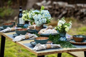 the photograph shows a picture from the side of a table set up outside filled with clay coyote pieces. the table has been set up for a dinner service, with multiple dinner place settings of dinner plates and soup bowls all glazed in joes blue. there is a large vase with a blue bouquet of flowers resting in it, the vase is glazed in joes blue. there is a deep salad bowl glazed in joes blue with multiple wicker balls decoratively placed within it. the wicker balls range from brown, to half brown half white to solid white. there is another bowl filled with these wicker balls, but it is mostly blocked from view by a soup and chili bowl, not allowing for identification. on the left side of the tablescape there is a bottle of wine, resting in a clay coyote wine coaster also in joes blue. there is a clay coyote yunomi near the wine bottle, also glazed in joes blue. each place setting has a linen napkin with a woven wooden napkin ring. each napkin ring has a small purple flower tucked into it. the photograph was taken outside with white natural light.